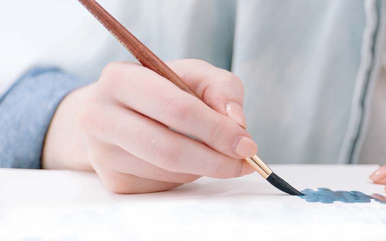 Una mano sujetando un pincel fino de madera, pintando sobre un papel.