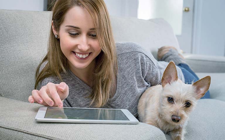 Una mujer recostada en un sillón sonríe mientras mira su dispositivo electrónico junto con su perro blanco.