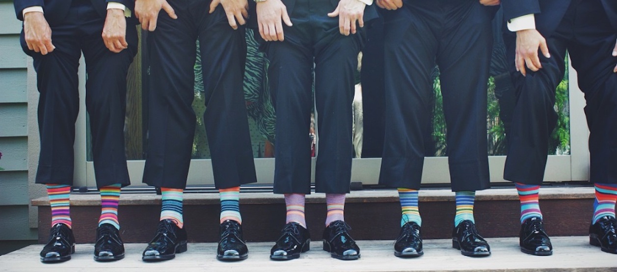 Piernas de personas que están levantando sus pantalones de traje de vestir negros para mostrar sus medias. Debajo de sus zapatos elegantes visten medias rayadas de muchos colores.