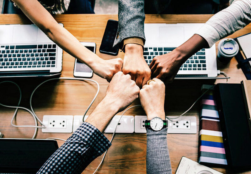 Cinco puños chocando sobre una mesa de trabajo, llena de cables, dispositivos electrónicos, agendas y café.